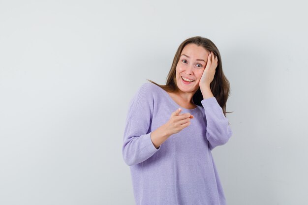 Young female in lilac blouse holding hand on her head while pointing forward and looking jolly 