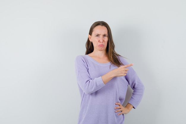 Young female in lilac blouse pointing aside and looking displeased 