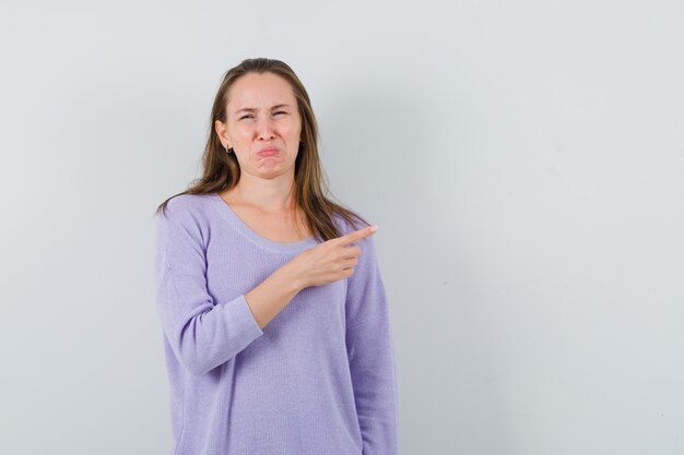 Young female in lilac blouse pointing aside and looking dissatisfied 