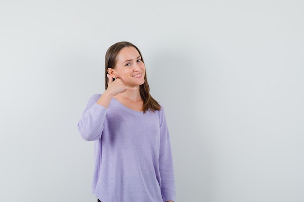 Молодая женщина в сиреневой блузке показывает жест телефонного звонка и выглядит довольной