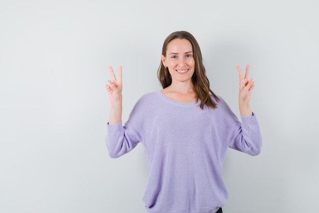 Young female in lilac blouse showing V-sign while smiling and looking optimistic 