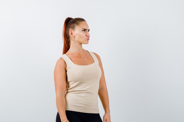 Young female looking away while curving lips in beige tank top and looking thoughtful. front view.