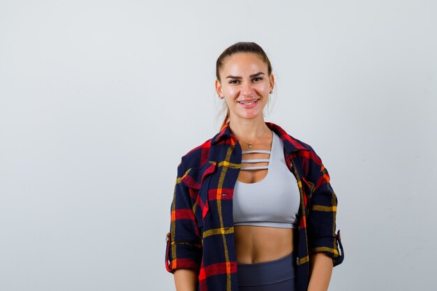Young female looking at camera in crop top, checkered shirt, pants and looking happy , front view.