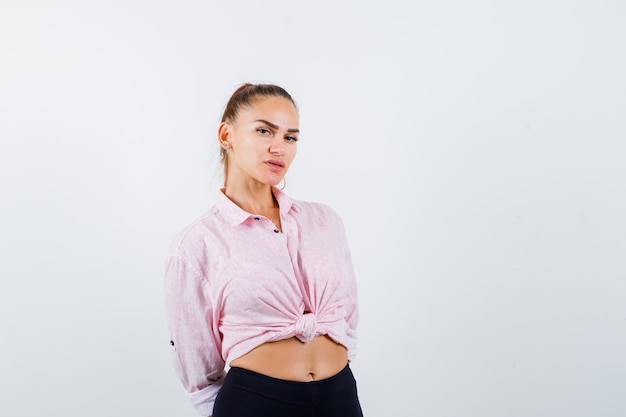 Free photo young female looking at camera while holding hands behind back in casual shirt, pants and looking confident. front view.