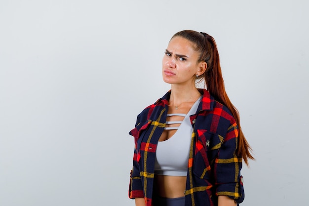 Young female looking at camera while scowling in crop top, checkered shirt, pants and looking serious , front view.