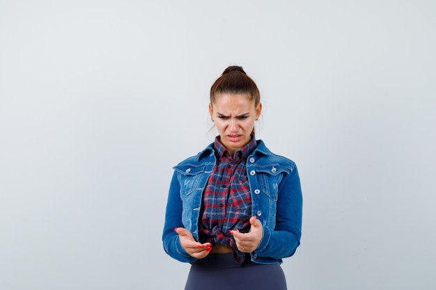 Young female looking down in checkered shirt, jacket, pants and looking indecisive. front view.