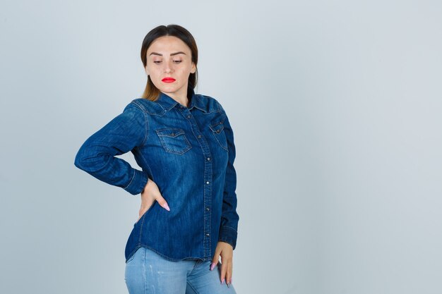 Young female looking down while keeping hand on hip in denim shirt and jeans and looking confident