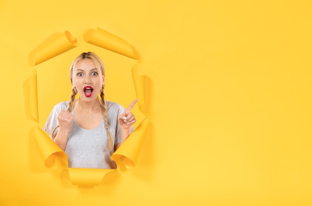 Free photo young female looking from the torn yellow paper background facial window indoor sale