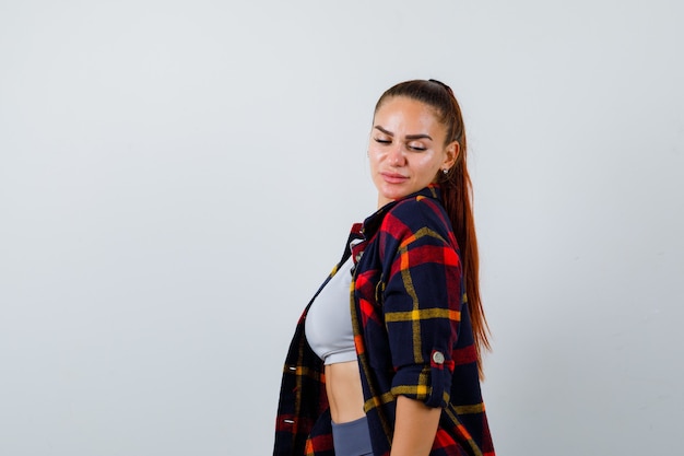 Young female looking over shoulder in crop top, checkered shirt, pants and looking pretty. front view.