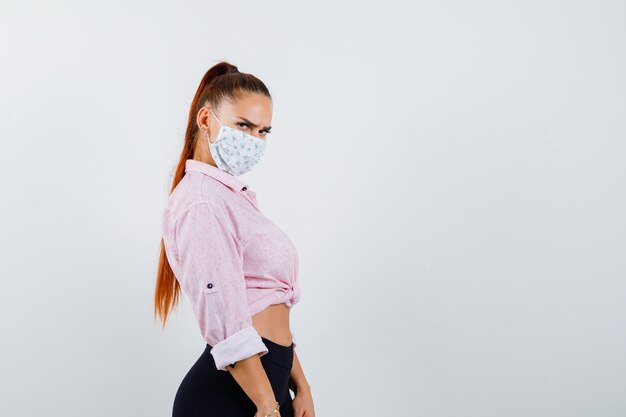 Молодая женщина смотрит через плечо в рубашке, штанах, медицинской маске и выглядит уверенно, вид спереди.
