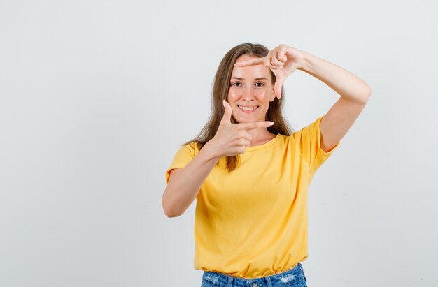 Young female making frame with fingers in t-shirt, shorts and looking cheery