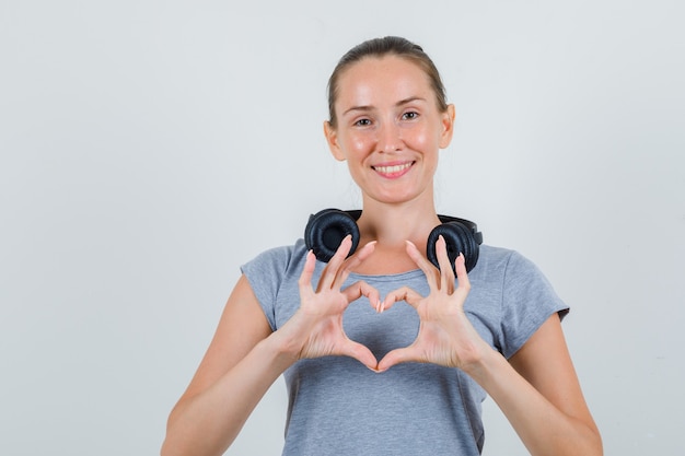 Free photo young female making heart shape with fingers in grey t-shirt and looking cheerful. front view.