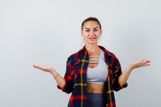 Young female making scales gesture in crop top, checkered shirt, pants and looking confident , front view.