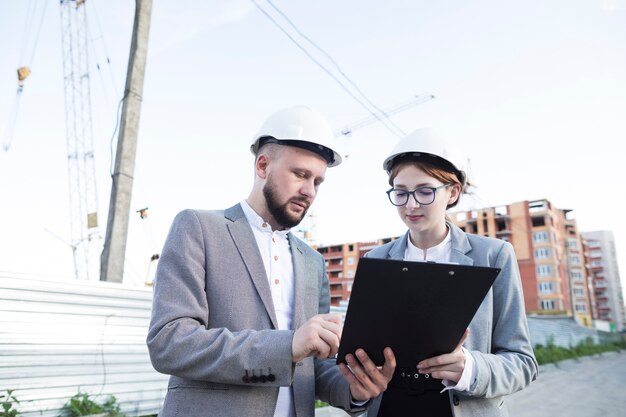 Young female and male architect wearing hard hat looking at clipboard