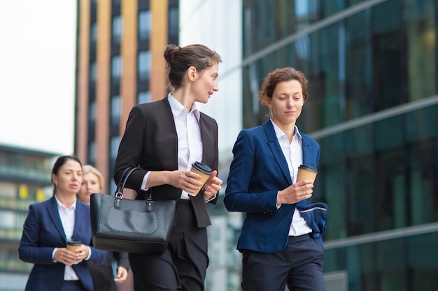 Young female managers with takeaway coffee mugs wearing office suits, walking together in city, talking, discussing project or chatting. Medium shot. Work break concept