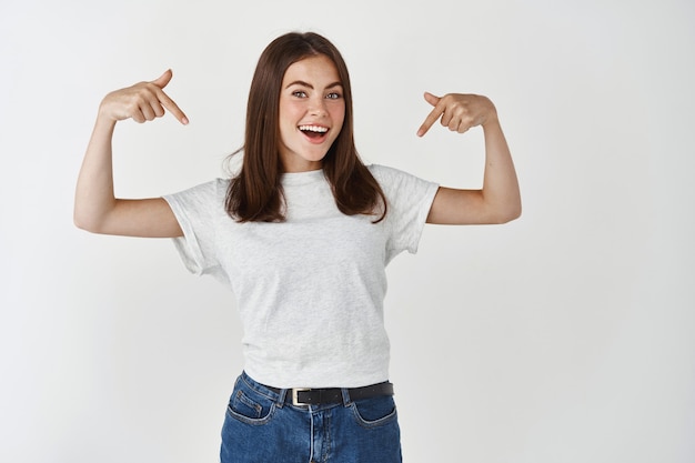 Young female model showing advertisement, pointing fingers down at t-shirt logo and smiling, standing over white wall.