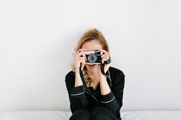 Молодая женщина-модель со светлыми волосами, одетая в черные пижамы, сидит над белой стеной и фотографирует на ретро-камеру, место для текста
