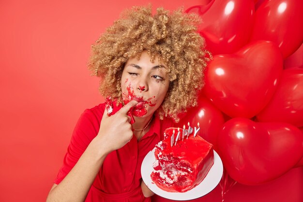 Young female model with curly hair licks finger has sweet tooth enjoys eating tasty cake smeared with cream wears dress poses near bunch of inflated heart balloons isolated over red background
