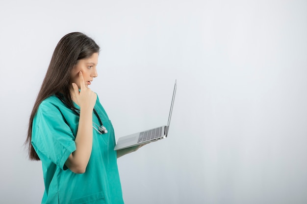 Young female nurse looking at something on laptop over white wall. 