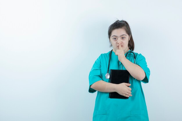 Young female nurse with stethoscope and notepad showing silent sign against white wall .