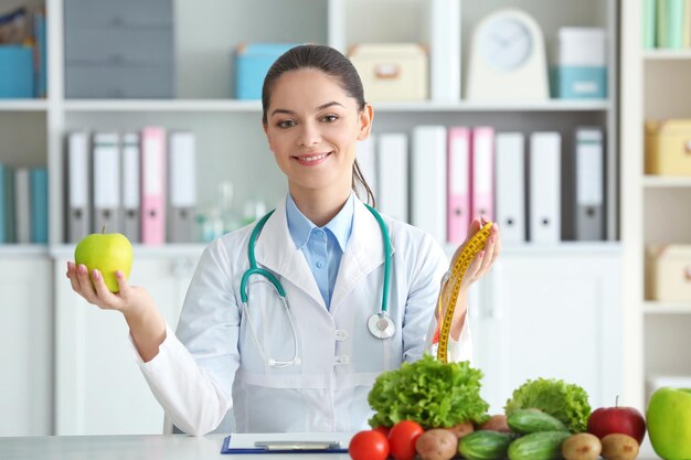 Young female nutritionist holding measuring tape and apple while sitting at table in her office