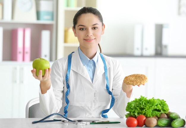 Young female nutritionist holding tasty cookie and apple while sitting at table in her office