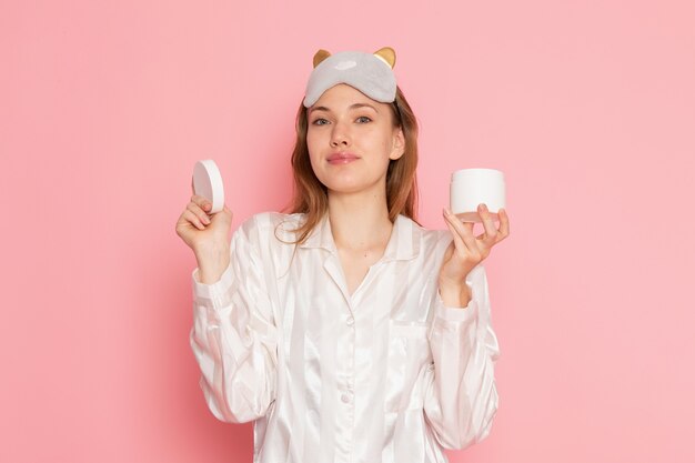 young female in pajamas and sleep mask holding cream and smiling on pink