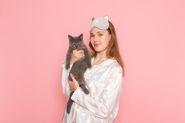 young female in pajamas and sleep mask holding cute grey cat on pink