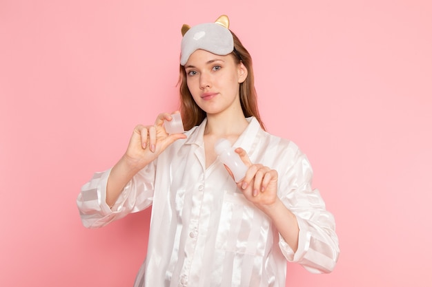 молодая женщина в пижаме и маске для сна держит средство для макияжа на розовом