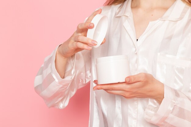 young female in pajamas and sleep mask opening cream on pink