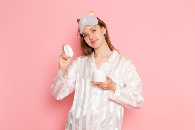 young female in pajamas and sleep mask posing with smile holding cream on pink