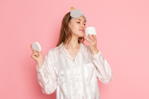 young female in pajamas and sleep mask smelling cream on pink