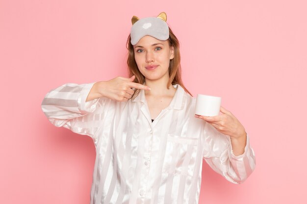 молодая женщина в пижаме и маске для сна улыбается, держит белую кремовую банку на розовом