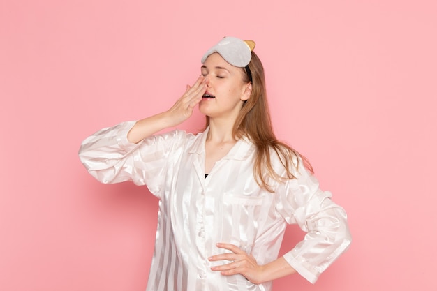 young female in pajamas and sleep mask yawning badly on pink