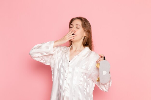 young female in pajamas and sleep mask yawning on pink