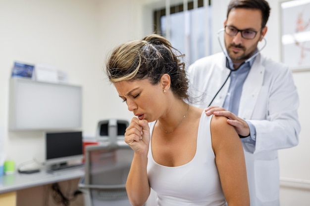 Young female patient in the clinic suffered from pneumonia she is coughing the doctor listens to the wheezing in the lungs with a stethoscope