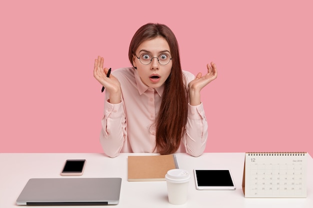 Young female perfectionist arranges all things neatly, has stupefied expression, stretches hands, surrounded with laptop computer, tablet, calender