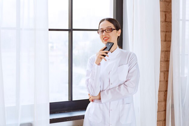 窓の近くに立っている間携帯電話を保持している白衣の若い女性医師。