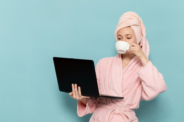 young female in pink bathrobe after shower drinking coffee and using laptop on blue