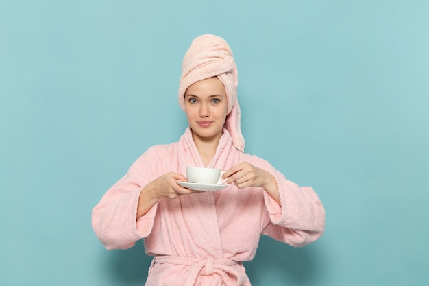 young female in pink bathrobe after shower drinking coffee with smile on blue