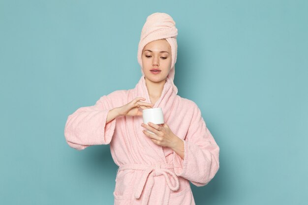 young female in pink bathrobe after shower holding cream using it on blue