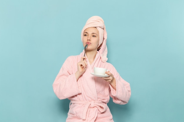 young female in pink bathrobe after shower holding cup of coffee on blue desk