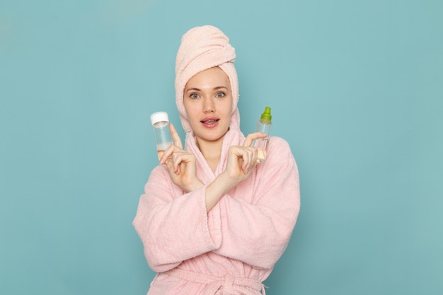 young female in pink bathrobe after shower holding different sprays on blue