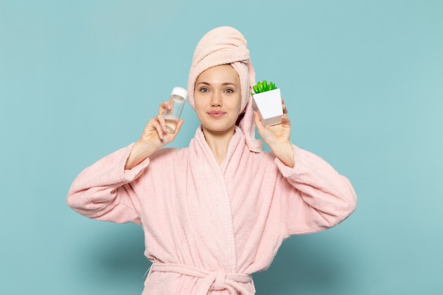 young female in pink bathrobe after shower holding green plant spray on blue