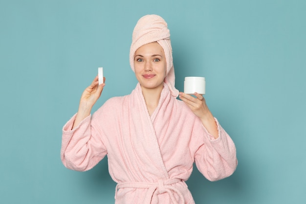 young female in pink bathrobe after shower holding spray and cream on blue