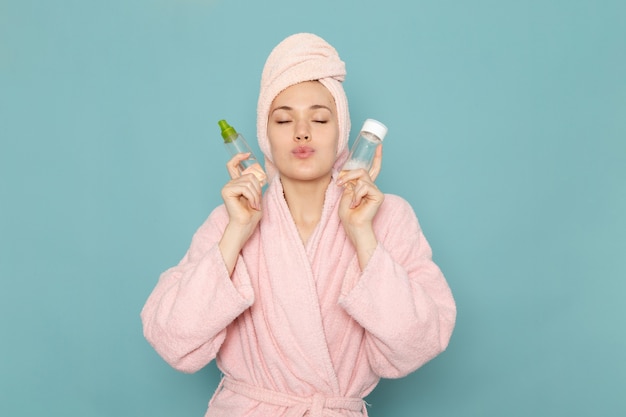 young female in pink bathrobe after shower holding sprays on blue