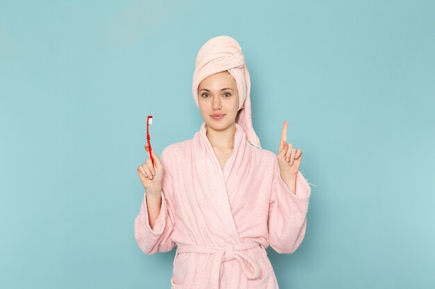 young female in pink bathrobe after shower holding toothbrush on blue