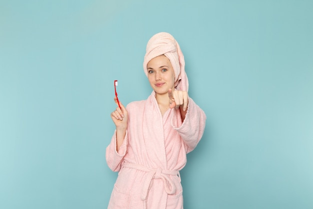 young female in pink bathrobe after shower holding toothbrush and smiling on blue