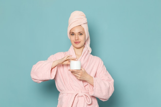 young female in pink bathrobe after shower holding white cream on blue