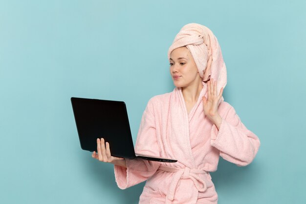 young female in pink bathrobe after shower using laptop on blue